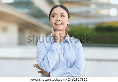A professional Chinese woman with her chin resting on her hand stands outdoors, looking up and away as if deep in thought. She wears a crisp blue shirt, her hair is neatly tied back