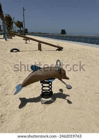 A children's playground features a dolphin-shaped spring toy on a sandy area with a beachside walkway and palm trees in the background