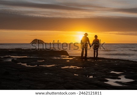 Couple holding hands and enjoying the sunset at Muriwai beach. Oaia Island in the distance. Auckland.  Royalty-Free Stock Photo #2462121811