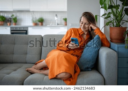Smiling woman using phone on sofa at home. Relaxed female enjoying comfort, chatting with friends on social networks, watch movies, series, play online games on smartphone. Slow life concept Royalty-Free Stock Photo #2462134683