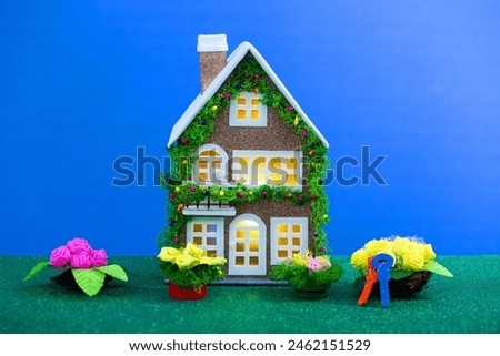 On a blue background, a brown house decorated with New Year's artificial garland with yellow lights in the windows and keys near the flowers