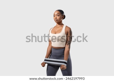 A young African American woman stands with poise, showcasing her athletic physique in a beige crop top and blue leggings with a striped resistance band, looking at copy space