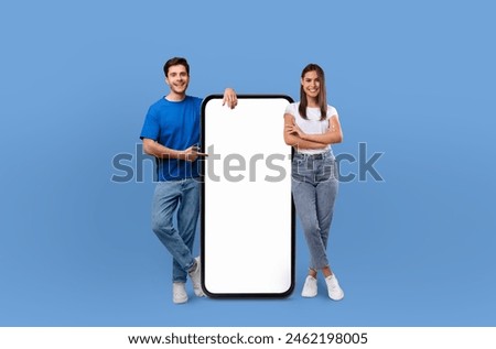 A cheerful young couple standing side by side, confidently presenting a large, blank mobile phone screen on a solid blue background.