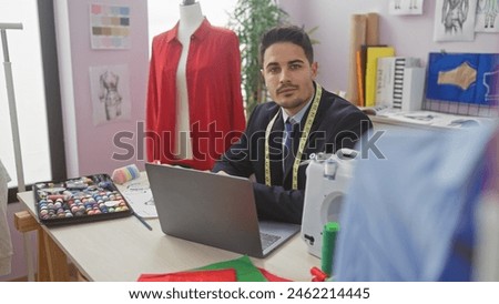 A handsome hispanic man with a beard works on a laptop in a colorful tailor shop surrounded by fashion designs and sewing materials.