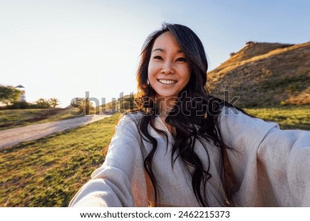 A young attractive Asian woman in a linen dress takes a selfie and laughs outdoors. Smiling Korean woman having fun and taking pictures in nature. Beautiful scenery and sunset.