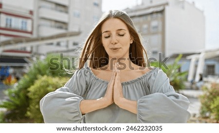 A serene young woman in a casual dress meditates with closed eyes, nestled amidst urban greenery.