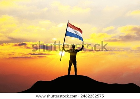 Paraguay flag being waved by a man celebrating success at the top of a mountain against sunset or sunrise. Paraguay flag for Independence Day.