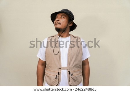 Macho mature man with beard and mustache wearing safari outfit isolated on beige background. Half body portrait of an adult Southeast Asian man posing wearing a vest, bucket hat and hip flask bottle
