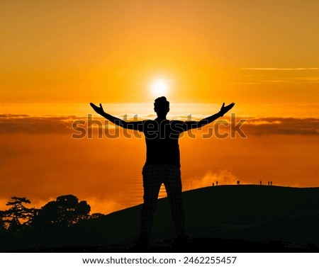 A man standing on highest place with open arms facing the sun when its setting, picture from backside made an excellent combination of orange and black. The posture gives the impression of motivation