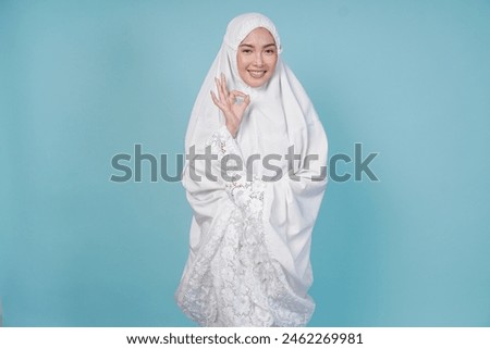 Young Asian Muslim woman wearing prayer gown showing okay sign with right hand, isolated by blue background. Hajj concept.