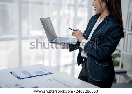 A woman in a business suit is holding a laptop and a pen, smiling as she presents something on the screen. Concept of professionalism and confidence, as the woman is well-prepared
