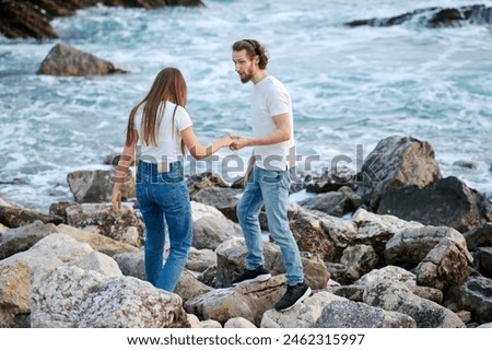 A man and a woman are standing on rocks near the ocean, looking out towards the water. The scene shows the couple enjoying the seaside view on a clear day.