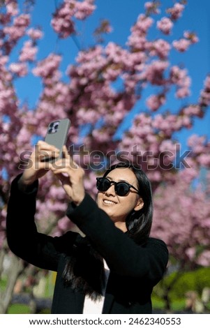 An Asian woman captures the beauty of blooming sakura with her smartphone camera in the park.
