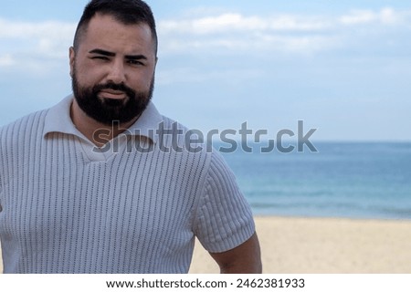 The image captures a 30-year-old man in a moment of serene contemplation by the sea. Dressed in a white knitted polo shirt, he exudes a relaxed elegance against the soothing backdrop of the ocean’s 