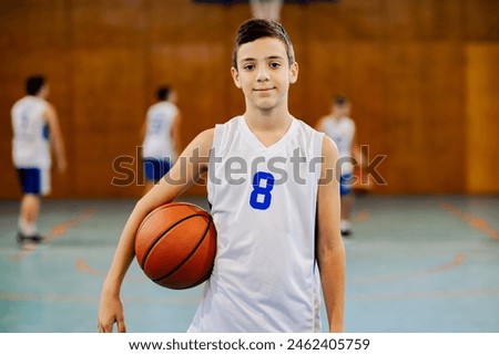 Portrait of a junior sporty athlete staining at indoor basketball court with a ball and smiling at the camera. Young basketball player posing at indoor court with a basketball and looking at camera.