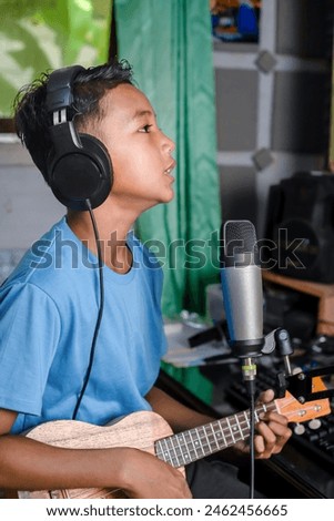 A child is recording a song in a home recording studio. A child playing the ukulele sings into a recording microphone