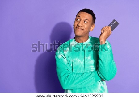 Photo of handsome thoughtful guy wear teal sweatshirt rising bank card looking emtpy space isolated purple color background
