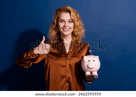 Portrait of satisfied woman with wavy hair dressed silk shirt hold money piggy box show thumb up isolated on dark blue color background
