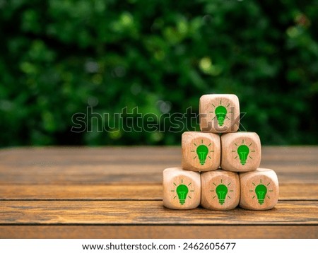 Green energy, creative idea thinking strategy, solution and eco innovation concept. Green light bulbs icon on wooden cube blocks pyramid stack on wood desk on nature garden background with copy space.
