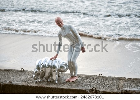 Young hairless girl with alopecia in white futuristic suit walking on concrete fence with toy tardigrade on sea background, bald pretty girl symbolizes courage and acceptance of unique appearance