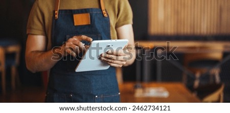 A male waiter in an apron uses a digital tablet, efficiently managing orders in a well-appointed bar, his efficient demeanor enhancing the customer experience.