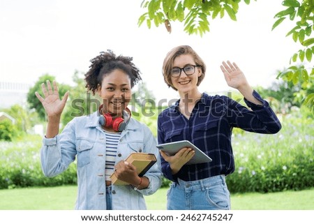 Two women are standing in a park, one of them is holding a book and the other is holding a tablet. They are both smiling and waving at the camera
