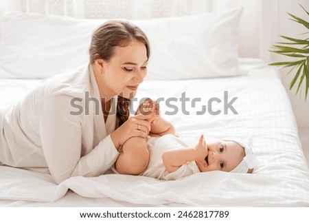 mom and baby play and rejoice in a bright white room on the bed, a mother with a small child hugs and kisses, happy motherhood with maternity leave, lifestyle