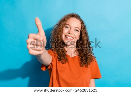 Portrait photo of youngster cheerful girl with beautiful curly red hair wearing orange t shirt makes high rating isolated on blue color background