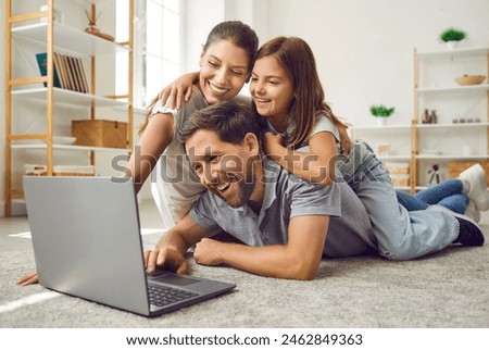 Portrait of happy cheerful young family with child girl lying on the floor at home using laptop. Smiling parents enjoying weekend together watching funny cartoon online or talking on video call.