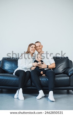 A young married couple with a small child is photographed sitting on a sofa on a white background. Caring parents look at their baby while holding it in their arms.