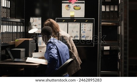 Multiethnic investigators examine evidence, discuss the case, and gather clues in a well-organized environment. Caucasian woman carrying forensic files joins african american man in incident room. Royalty-Free Stock Photo #2462895283