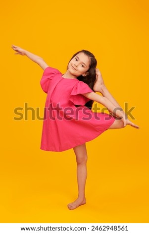 A joyful girl with long dark hair, dressed in a pink summer dress, standing on one leg in a swallow pose. Yellow background. Summer. Happy children.