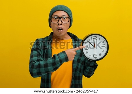 An excited young Asian man, dressed in a beanie hat and casual shirt, appears pleasantly surprised as he points to the clock he is holding while standing against a yellow background