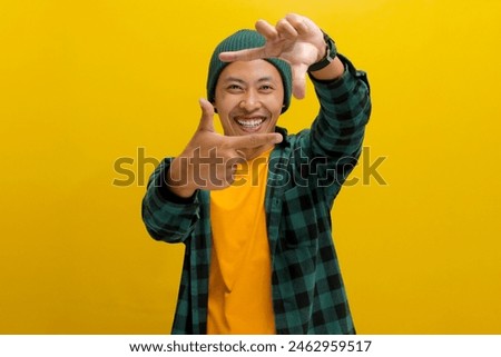 Happy young Asian man, filled with inspiration, is imagining how to capture an interesting shot. He's making a frame gesture with his hands and fingers, as if framing the composition for a photograph