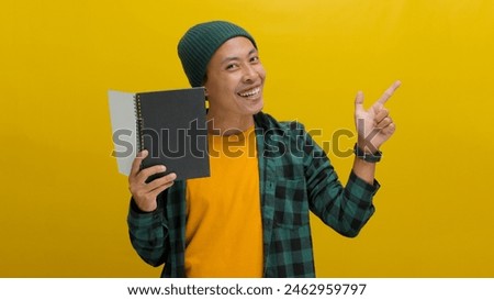 An excited young Asian student, dressed in a beanie hat and casual clothes, is smiling while pointing aside at an empty copy space, standing against a yellow background