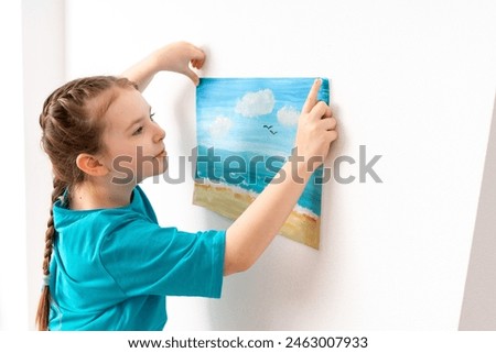 Cute little girl hanging her drawing on the wall. A girl places a seascape on the wall, dreaming of the sea. Summer seascape painted with paints. High quality photo