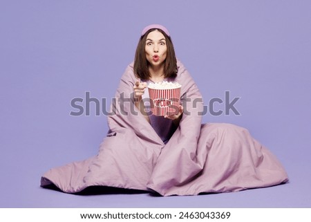 Full body young scared woman wears pyjamas jam sleep eye mask rest relax at home sit wrapped in blanket eat pop corn watch movie film isolated on plain purple background. Bad mood night nap concept