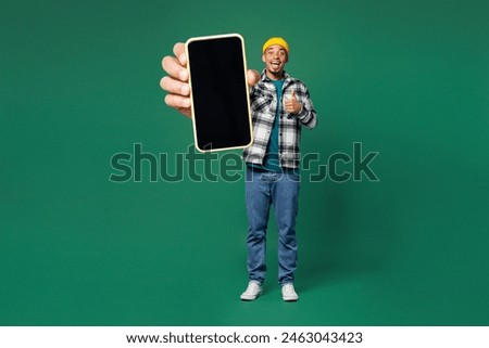Full body young man of African American ethnicity wear shirt blue t-shirt yellow hat hold in hand use blank screen mobile cell phone show thumb up isolated on plain green background. Lifestyle concept
