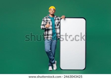 Full body young man he wears shirt blue t-shirt yellow hat big huge blank screen mobile cell phone smartphone with workspace area show thumb up isolated on plain green background. Lifestyle concept