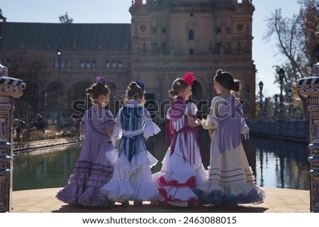 Four girls dancing flamenco, with their backs turned and talking to each other, in typical flamenco costumes next to a lake in a beautiful square in Seville. Concept dance, flamenco, typical Spanish. Royalty-Free Stock Photo #2463088015
