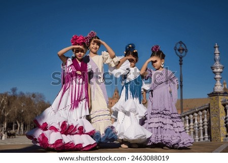 Four girls dancing flamenco, covering their eyes from the sun with their hands, in typical flamenco costumes on a bridge in a beautiful square in Seville. Dance concept, flamenco, typical Spanish. Royalty-Free Stock Photo #2463088019