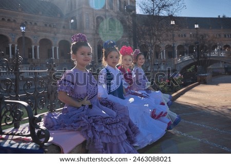 Four girls dancing flamenco, looking at camera, in typical flamenco costumes sitting on a bench in a nice square in Seville, receiving sun rays. Dance concept, flamenco, typical Spanish, Spain. Royalty-Free Stock Photo #2463088021