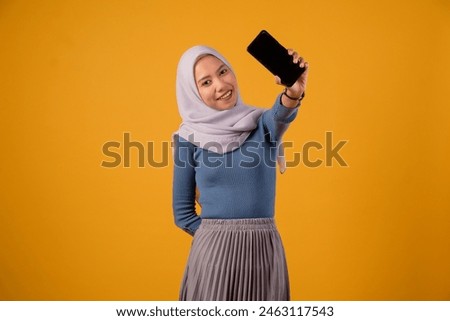 Portrait of smiling beautiful Asian woman in blue sweater and hijab using a mobile phone and looking aside isolated over yellow background