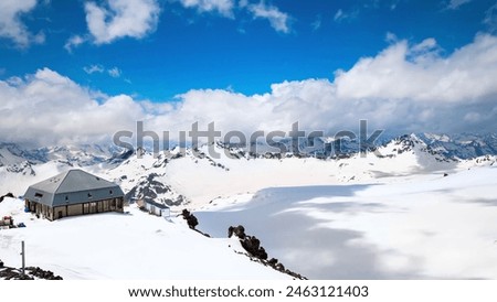 The snow-covered mountain peaks, the cozy cabin, and the clear blue sky create a serene and picturesque scene. Royalty-Free Stock Photo #2463121403
