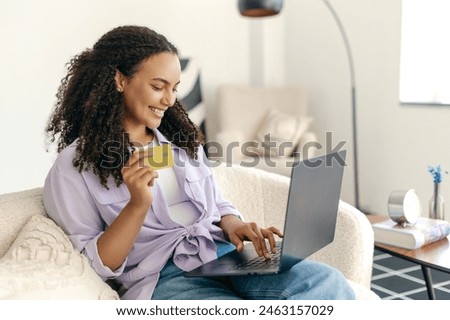 Online shopping. Positive hispanic or brazilian curly haired woman, using credit card and laptop to buying something, paying online making purchase in online store while sitting at home, smiling