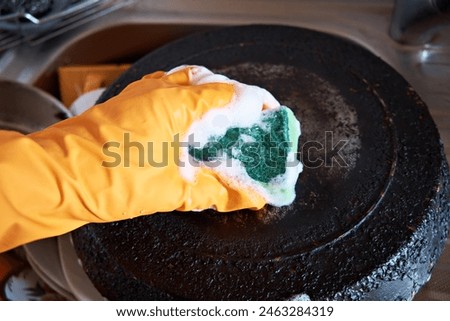 A man's hand in a protective glove cleans a pan with a burnt bottom with a foam kitchen sponge. Royalty-Free Stock Photo #2463284319