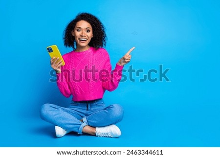 Full size photo of pretty young girl sit floor point empty space hold device wear trendy pink outfit isolated on blue color background