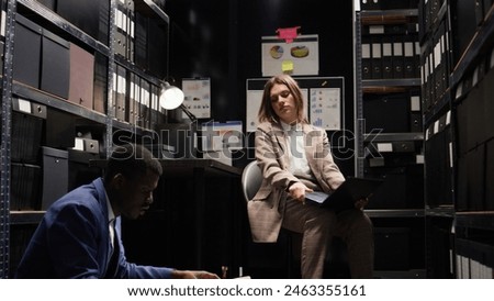In incident room, multiethnic team of weary private investigators analyzes evidence, reviews files, and does research to solve the case. Two detectives are exhausted by criminal investigation reports. Royalty-Free Stock Photo #2463355161