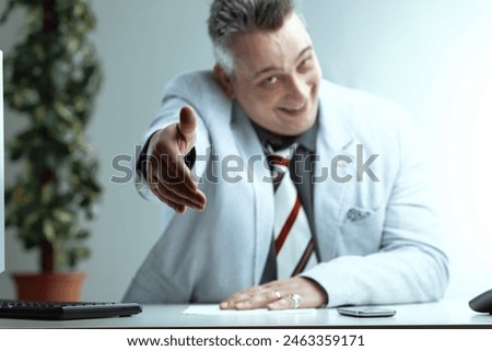 Man with spiky gray hair, light gray suit jacket, dark gray shirt, and striped tie, extends a friendly handshake with a cheerful expression, hiding deceitful intentions, seated at a desk Royalty-Free Stock Photo #2463359171