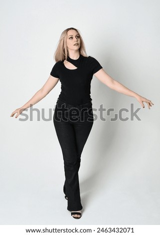 Full length portrait of beautiful blonde woman wearing modern black shirt and leather pants. Confident standing pose with gestural hands presenting, silhouetted on white studio background.